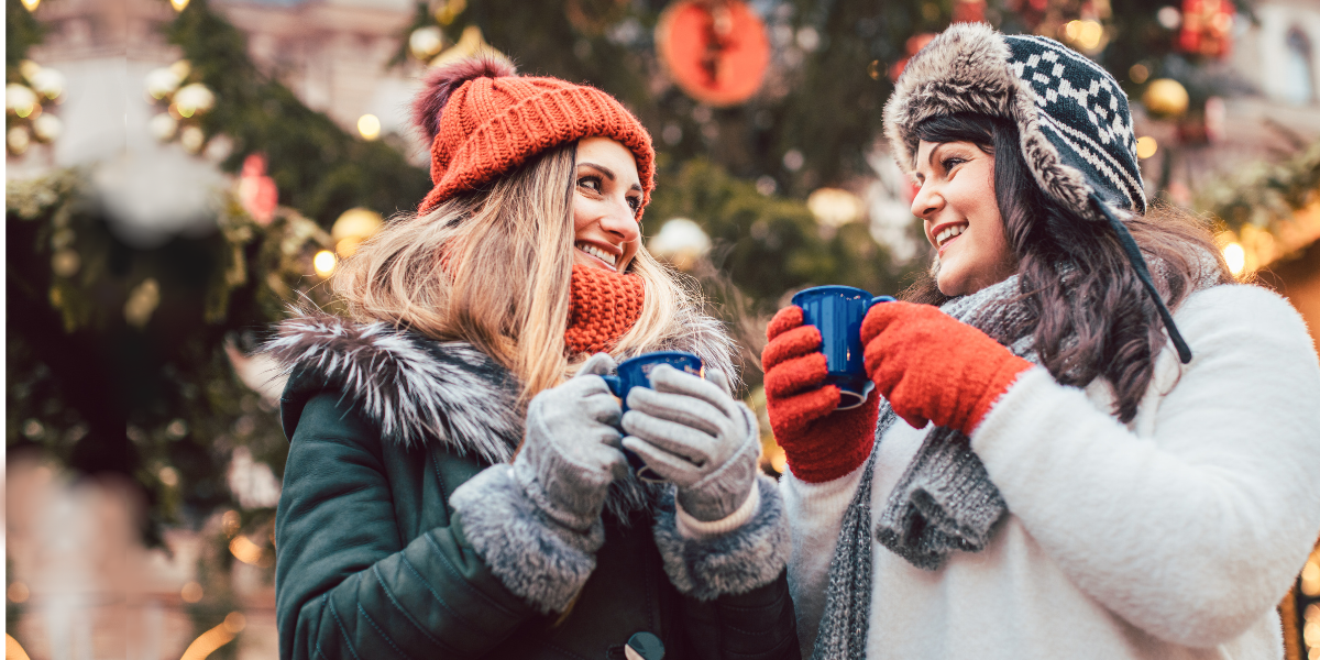 Two women who are friends walking outside during christmas. They have mugs of hot cocoa and are walking past a christmas tree