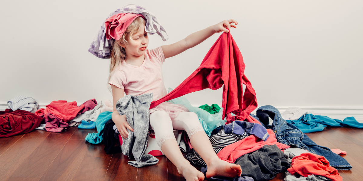 A little girl sitting a pile of clothing. She is perplexed by the large mess.