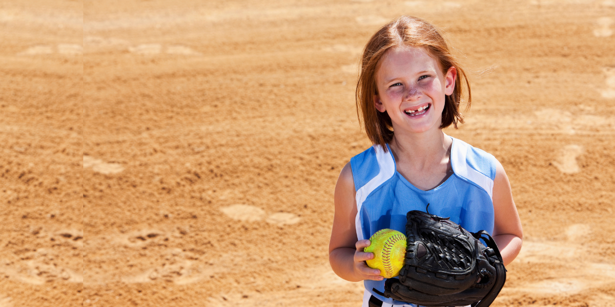 A young redheaded girl smiling at the camera as she prepares to play softball. She has a mitt on and is holding a softball in the other hand