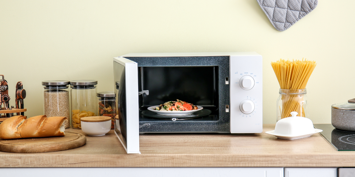 An open Microwave with re-heated pasta inside. Beside it on the counter, there are several jars, a butter dish, and some bread on a cutting board.