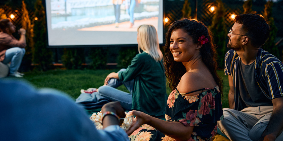 A woman and her friends during an outdoor movie night. She is handing her friend a bowl of popcorn