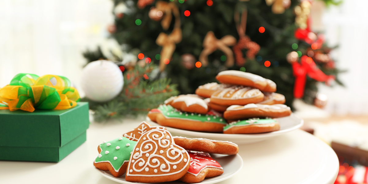 Two plates of Christmas cookies on a table. There is a gift behind them and a Christmas tree in the background