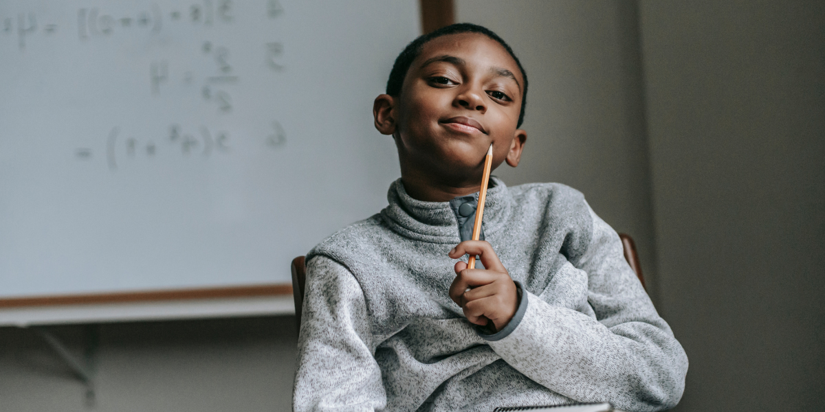 A little boy thinking deeply in a classroom. He's tapping a pencil on his chin.