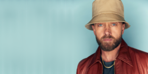 TobyMac wearing a tan bucket hat, a reddish-brown leather jacket, and a gold chain over a black t-shirt. The background is a pale blue gradient
